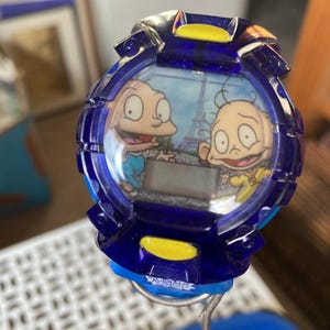 May include: A blue digital watch featuring cartoon characters. The watch face displays two characters with the Eiffel Tower in the background. The watch has a blue bezel with yellow accents.