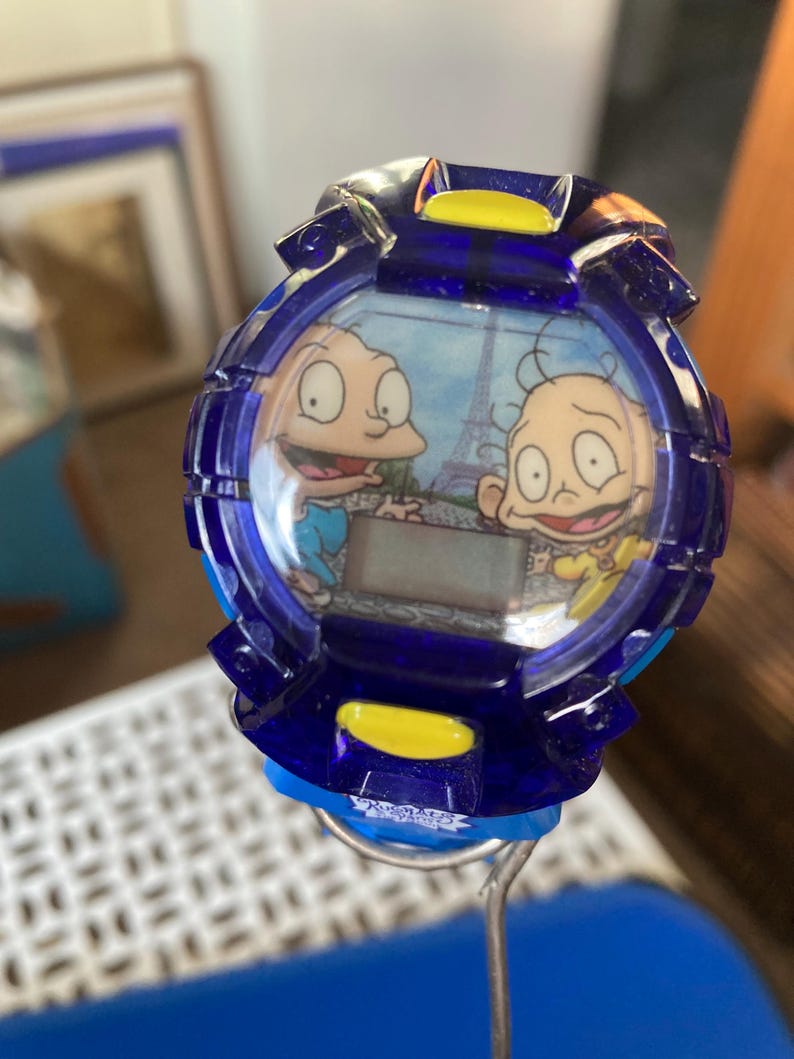 May include: A blue digital watch featuring cartoon characters. The watch face displays two characters with the Eiffel Tower in the background. The watch has a blue bezel with yellow accents.
