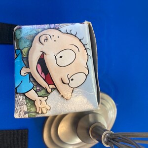 May include: A small, rectangular box featuring a cartoon image of Tommy Pickles from the animated series Rugrats. The box has a light blue background with a cartoon image of Tommy Pickles. The box is resting on a metal object.