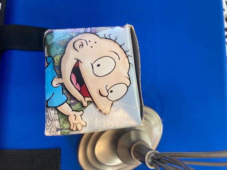 May include: A small, rectangular box featuring a cartoon image of Tommy Pickles from the animated series Rugrats. The box has a light blue background with a cartoon image of Tommy Pickles. The box is resting on a metal object.
