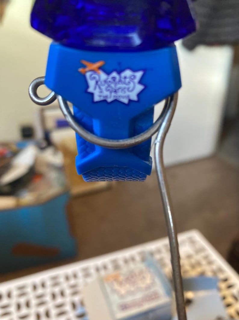 May include: A blue bottle stopper with a silver metal handle. The stopper has the text "Rugrats" in white script inside a starburst design. The top of the stopper is a dark blue, possibly glass.