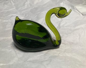 May include: A handcrafted glass swan figurine in shades of green. The swan has a curved neck and a rounded body. The glass has a glossy finish, and the figurine is sitting on a white surface. A decorative item.