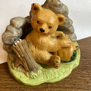 May include: A ceramic figurine of a brown bear sitting on a green base with a small pumpkin. The bear is surrounded by rocks and a tree stump.
