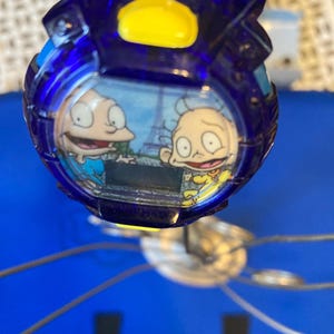 May include: A blue and yellow digital watch featuring cartoon characters. The watch face displays two characters with the Eiffel Tower in the background. The watch has a blue, transparent casing and a yellow button.