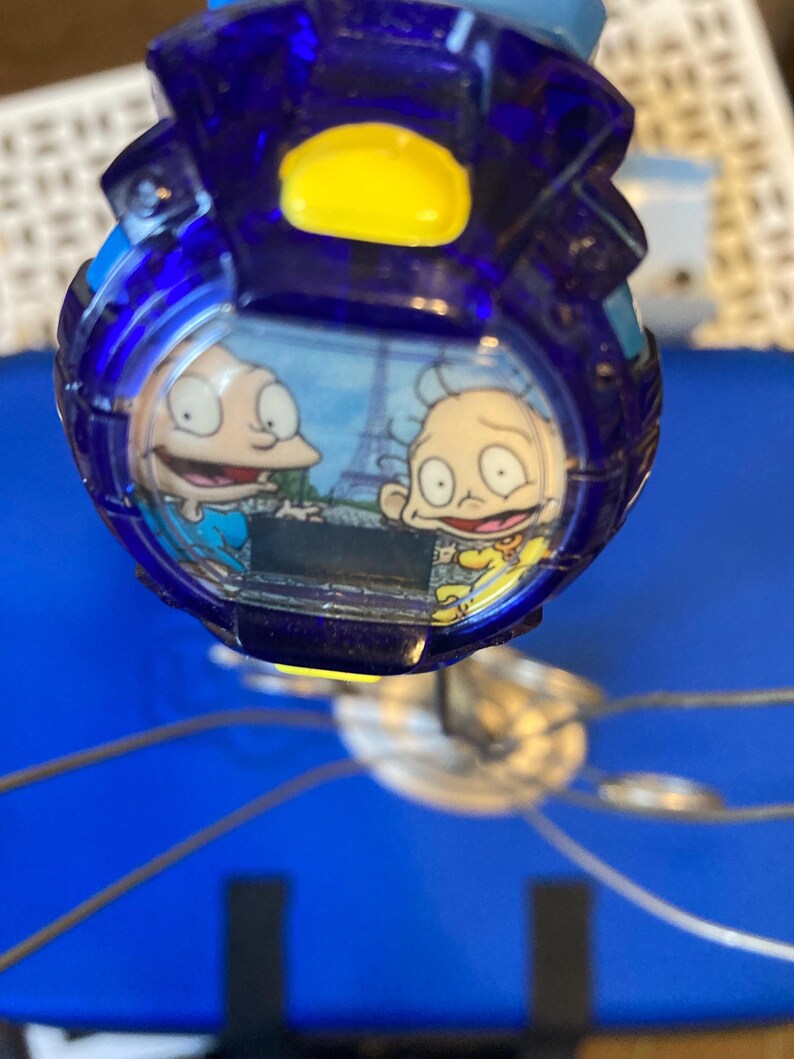 May include: A blue and yellow digital watch featuring cartoon characters. The watch face displays two characters with the Eiffel Tower in the background. The watch has a blue, transparent casing and a yellow button.