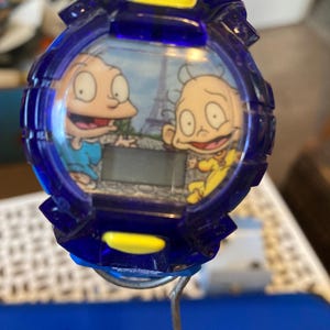 May include: A blue digital watch featuring cartoon characters Tommy and Chuckie from Rugrats, with the Eiffel Tower in the background. The watch has a blue bezel with yellow accents and a digital display.