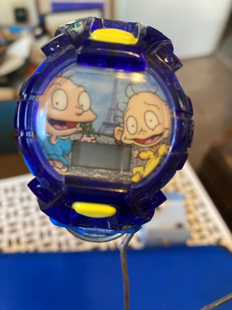 May include: A blue digital watch featuring cartoon characters Tommy and Chuckie from Rugrats, with the Eiffel Tower in the background. The watch has a blue bezel with yellow accents and a digital display.