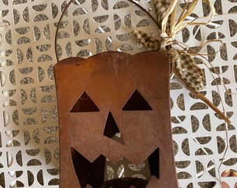 May include: A rusty metal jack-o'-lantern candle holder with a handle and a straw bow. The holder has a cut-out face with a wide smile, two triangle eyes, and a triangle nose.