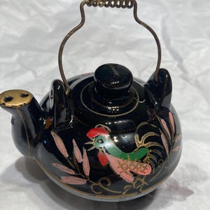 May include: Black ceramic teapot with a gold-colored handle and spout. The teapot is decorated with a rooster design in pink, green, and gold.