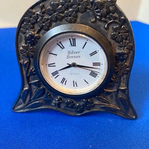 May include: A silver-toned metal table clock with an arched frame featuring embossed floral details. The white clock face displays black Roman numerals, the text Silver Scenes, and the word Quartz, with black hour, minute, and second hands.