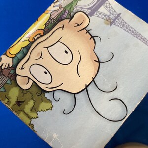 May include: A close-up of a cartoon illustration featuring a character's face with large eyes and curly hair. The background includes a depiction of the Eiffel Tower and a park scene, all on a light blue surface.
