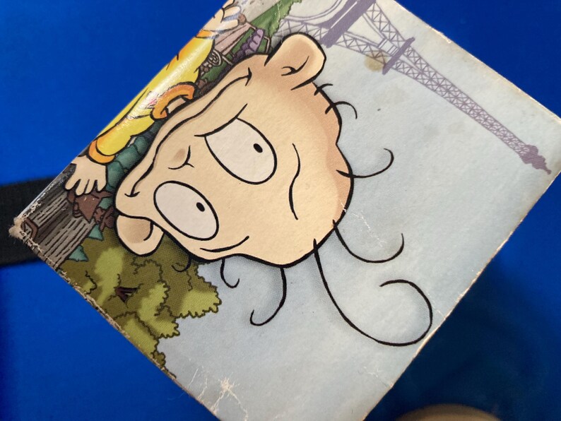 May include: A close-up of a cartoon illustration featuring a character's face with large eyes and curly hair. The background includes a depiction of the Eiffel Tower and a park scene, all on a light blue surface.