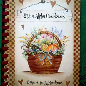 May include: A spiral-bound cookbook titled "Sigma Alpha Cookbook" with a decorative cover. The cover features a watercolor illustration of a basket overflowing with flowers, a pineapple, and a pumpkin. The words "Sisters in Agriculture" are also on the cover.