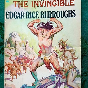 May include: Vintage paperback book cover for "Tarzan the Invincible" by Edgar Rice Burroughs. The cover art depicts Tarzan in a loincloth, with a woman and other figures, in a jungle setting. The title and author's name are prominently displayed.