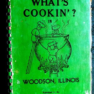 May include: A green cookbook with a spiral binding. The cover features a cartoon illustration of three chefs cooking in a large pot over a fire. The text on the cover reads "What's Cookin'? In Woodson, Illinois. Published by the American Legion Auxiliary."