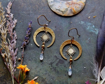 Könnte beinhalten: Ein Paar goldene Ohrringe mit einem Halbmond-Design und einem kleinen Sonnen-Anhänger, der an jedem Ohrring hängt. Die Ohrringe haben einen klaren Kristalltropfen, der an der Unterseite hängt.