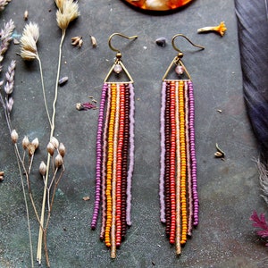 Könnte beinhalten: Ein Paar lange, geperlte Ohrringe mit einem dreieckigen Oberteil und einem Fransen aus bunten Perlen in den Farben Lila, Orange und Braun.