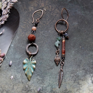 Könnte beinhalten: Ein Paar handgefertigte Ohrringe im Boho-Stil. Ein Ohrring hat einen Kupfer-Blatt-Anhänger, einen Kupferring, eine braune Perle und einen Kupferhaken. Der andere Ohrring hat einen Kupfer-Feder-Anhänger, einen Kupfer-Diamant-Anhänger, einen Kupferring, einen Kupferstab, grüne Perlen und einen Kupferhaken.
