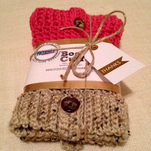 May include: A pair of handmade crochet boot cuffs in pink and beige, secured with brown buttons and twine. A tag with the word "THANKS" is attached. The cuffs are wrapped with a brown ribbon.