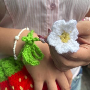以下が含まれることがあります： 鮮やかな赤色の本体に黄色の種のようなアクセント、緑色の葉のトップが付いたイチゴ型のクロシェ編みポーチ。緑色の茎には黄色い中心を持つ小さな白い花が取り付けられており、白いビーズのブレスレットが添えられています。