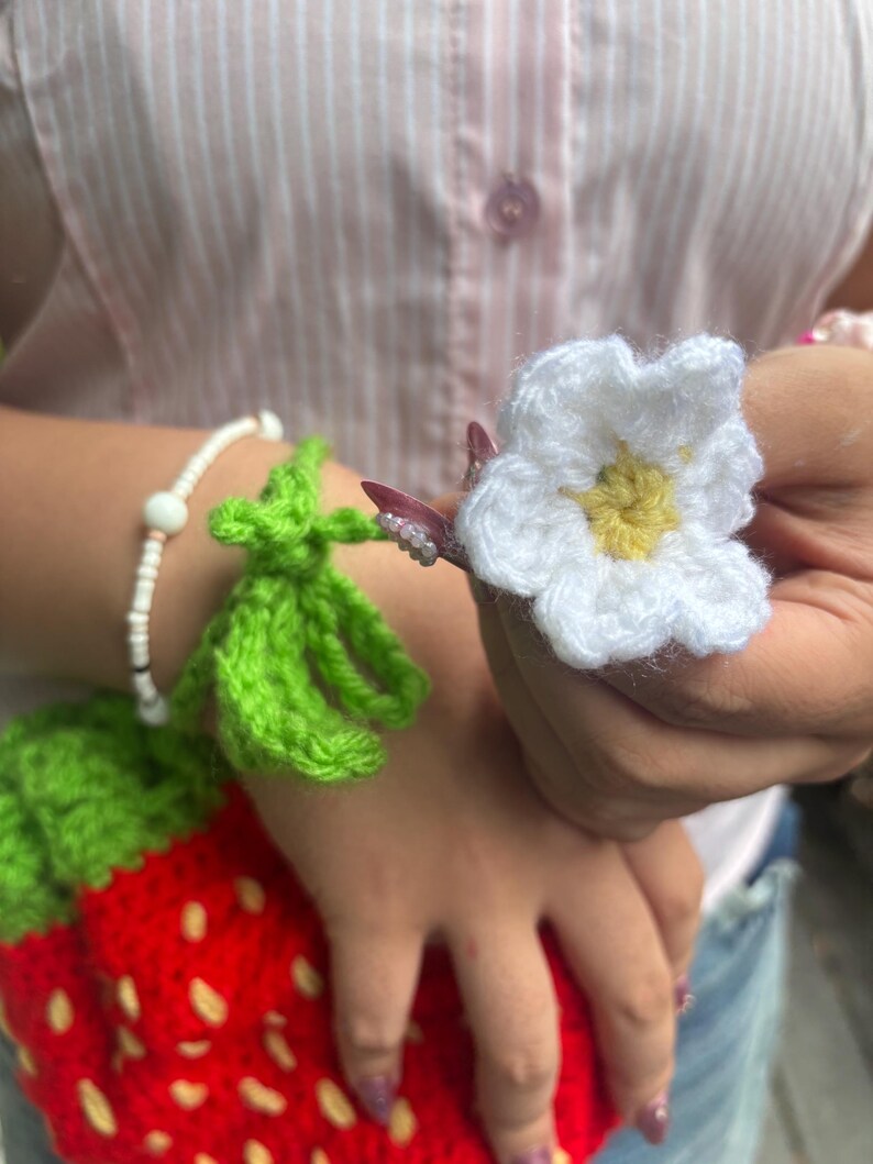 以下が含まれることがあります： 鮮やかな赤色の本体に黄色の種のようなアクセント、緑色の葉のトップが付いたイチゴ型のクロシェ編みポーチ。緑色の茎には黄色い中心を持つ小さな白い花が取り付けられており、白いビーズのブレスレットが添えられています。