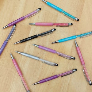 May include: An assortment of personalized pens in various colors, including purple, pink, silver, black, and teal. Each pen features a crystal-filled barrel and a name engraved on the side. The pens have a stylus tip and are arranged on a wooden surface.