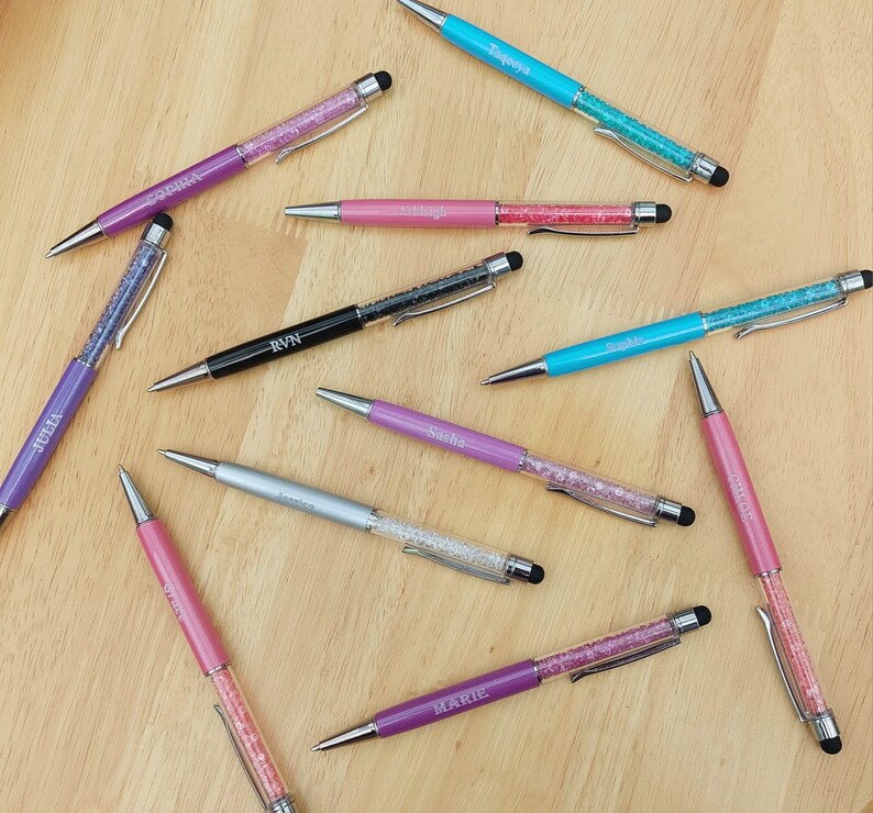 May include: An assortment of personalized pens in various colors, including purple, pink, silver, black, and teal. Each pen features a crystal-filled barrel and a name engraved on the side. The pens have a stylus tip and are arranged on a wooden surface.