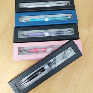 May include: A collection of personalized pens in various colors, each displayed in a black or colored gift box. The pens have crystal accents and are engraved with names like "Tageeya", "Julia", and "Chloe".