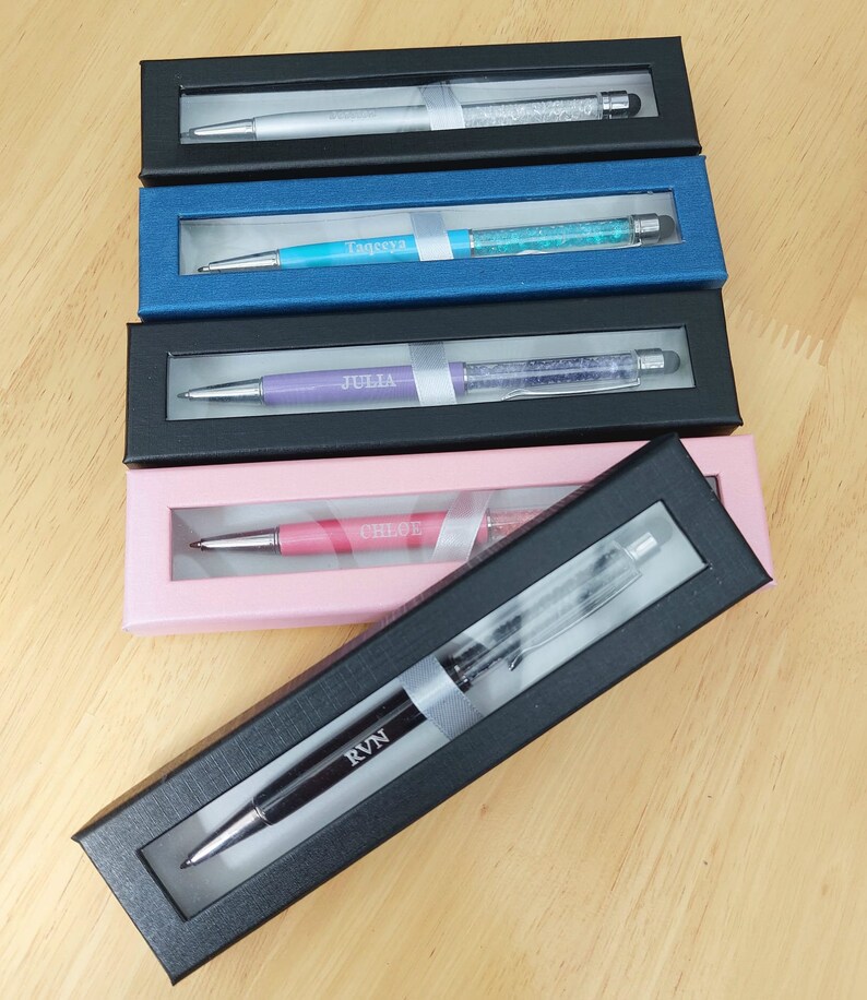 May include: A collection of personalized pens in various colors, each displayed in a black or colored gift box. The pens have crystal accents and are engraved with names like "Tageeya", "Julia", and "Chloe".