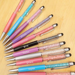May include: A collection of personalized pens with crystal-filled barrels and stylus tips. The pens feature various colors, including pink, blue, purple, and black, with names like "Amy," "Sophia," and "Julia" engraved on them. The pens are arranged on a light wooden surface.