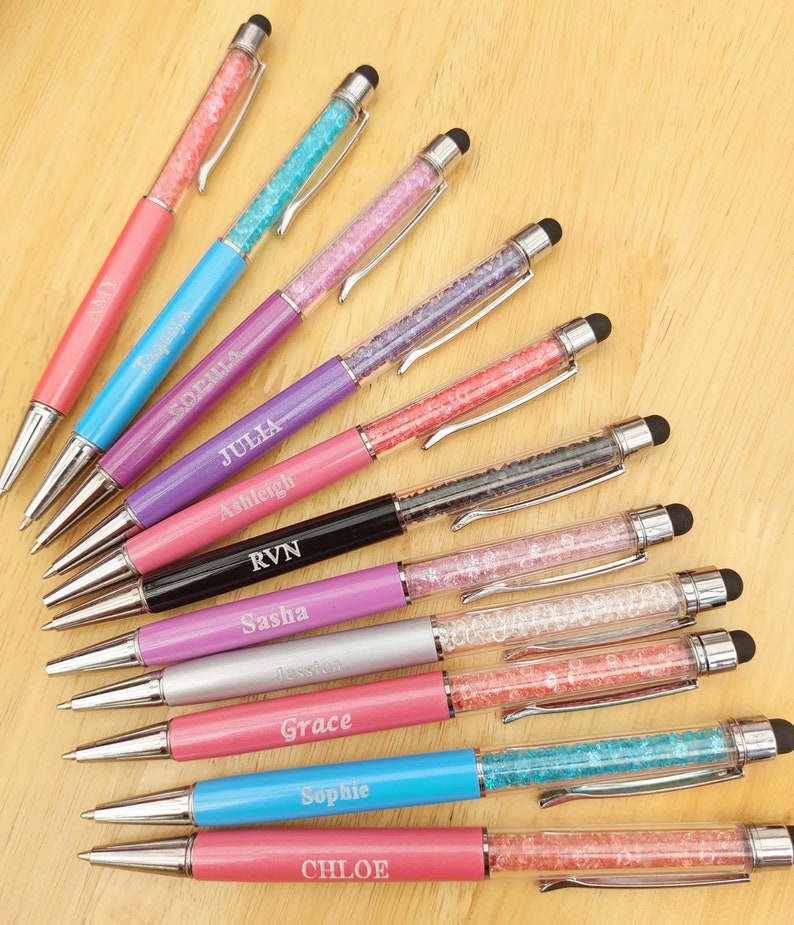 May include: A collection of personalized pens with crystal-filled barrels and stylus tips. The pens feature various colors, including pink, blue, purple, and black, with names like "Amy," "Sophia," and "Julia" engraved on them. The pens are arranged on a light wooden surface.