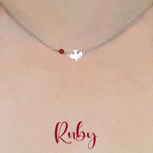 May include: A silver necklace with a small dove charm and a single red ruby bead. The necklace is on a model's neck and the word "Ruby" is written in red below the necklace.