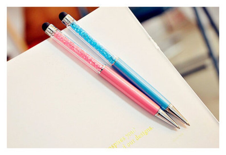 May include: Two pens with a stylus tip, one pink and one blue, are displayed on a white surface. The upper barrel of each pen is filled with sparkling crystals. The pens have silver-colored metal accents.