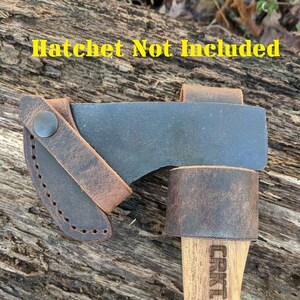 May include: A brown leather sheath with a snap closure for a hatchet. The sheath is attached to a wooden handle with the word "CRKT" engraved on it. "Hatchet Not Included" is written in yellow text at the top of the image.