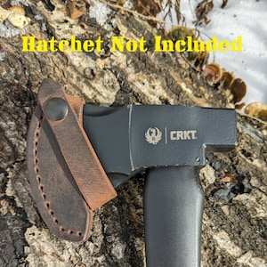 May include: A brown leather sheath with a snap closure for a hatchet. The sheath is attached to a black hatchet with a black handle and a silver CRKT logo. The text "Hatchet Not Included" is visible at the top of the image.
