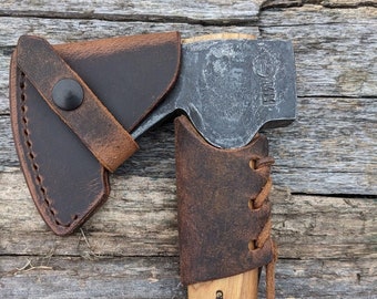 May include: A brown leather sheath with a snap closure for a small axe. The axe head is made of steel and has a wooden handle with the text "Gränsfors Bruk Sweden" printed on it. The text "Hatchet Not Included" is printed in red at the top of the image.