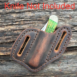 May include: A brown leather knife sheath with a stitched border and two belt loops. The sheath holds a knife with a green and white handle. The text "Knife Not Included" is visible in red.