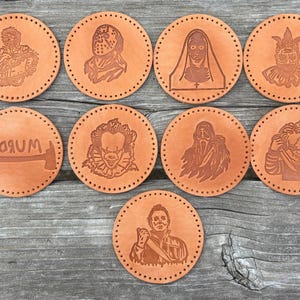 May include: A collection of ten round, tan leather coasters. Each coaster features a detailed, engraved image of a horror movie character. The images include a chainsaw-wielding figure, a masked killer, and a clown.