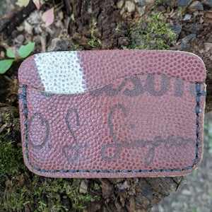 May include: A brown leather cardholder with a football texture and white stripes. The cardholder has black stitching around the edges and a handwritten signature. The cardholder is a unique accessory.
