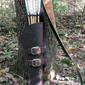 May include: A brown leather quiver with two silver buckles holds a set of arrows with white fletching. A wooden bow with green accents is leaning against a tree trunk.