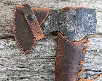 May include: A brown leather axe sheath with a lace-up strap and a brown wooden handle. The text "GRANSFORS BRUK SWEDEN" is printed on the handle. The text "Hatchet Not Included" is printed above the axe sheath.
