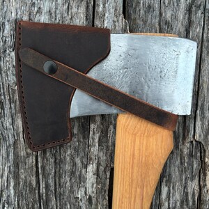 May include: A brown leather axe sheath with a strap and black button closure. The sheath covers the blade of an axe with a wooden handle. The text "AXE NOT INCLUDED" is in yellow letters at the top.