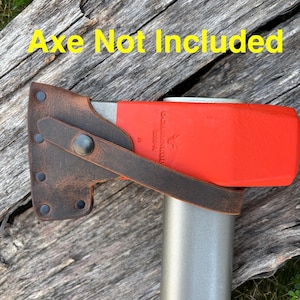 May include: Axe head with a red blade and a brown leather sheath. The sheath is secured with a strap and rivets. The axe is resting on a weathered wooden surface. Text on the image reads "Axe Not Included."