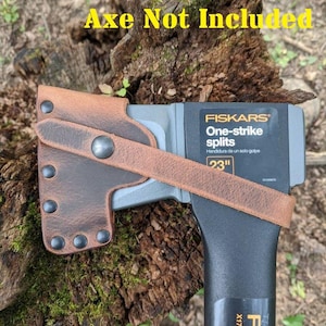 May include: A brown leather axe sheath with rivets and a strap, designed to fit a Fiskars X17 splitting axe. The axe is black and gray, with the text "FISKARS One-strike splits" and "23" visible.
