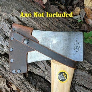 May include: A brown leather axe sheath with a black strap and rivets. The sheath is attached to a wooden axe handle with a yellow sticker that says "100% Made in USA". The axe head is silver and has the text "GRAHAM & NEVILLE USA Est. 1864" on the side. The text "Axe Not Included" is written in yellow letters on a brown wooden background.