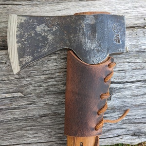 May include: A brown leather sheath with a brown leather strap and laces. The sheath is wrapped around a wooden handle with the text "Gränsfors Bruk Sweden" printed on it. The text "Hatchet Not Included" is printed in yellow on a wooden surface behind the sheath.