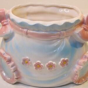 May include: A white ceramic planter shaped like a dress with pink bows and blue and white ruffles. The planter has a light blue glaze and is decorated with pink flowers.