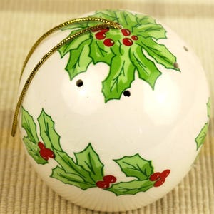 以下が含まれることがあります： 白いセラミック製のクリスマスオーナメント。ヒイラギのデザインで、緑色のヒイラギの葉と赤い実が特徴です。吊り下げ用の金色の紐が付いています。オーナメントは丸く、小さな穴が3つあります。