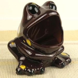 May include: A ceramic frog figurine in a glossy, dark brown finish with yellow accents. The frog has large, black eyes and an open mouth, suggesting it could be a container or a decorative piece. The figurine is on a light-colored surface.
