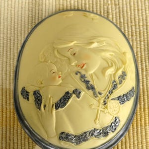 May include: Cream-coloured oval relief sculpture of a mother and child. The mother, with flowing hair, cradles the baby. The relief has a silver-coloured metal frame and decorative blue and silver accents.